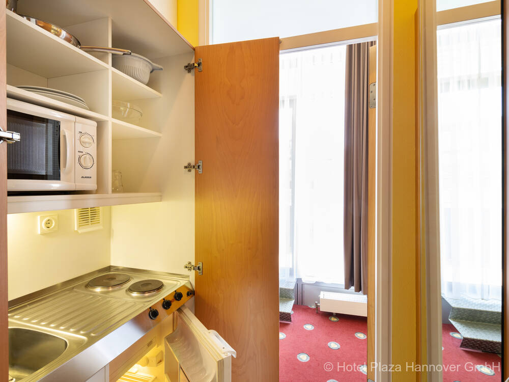 Kitchenette im Schrank mit Holztüren und weißer Küchenausstattung, inklusive Mikrowelle, Spüle, Herd, Kühlschrank und Zubehör. Bodentiefe Fenster lassen viel Licht in den Raum.